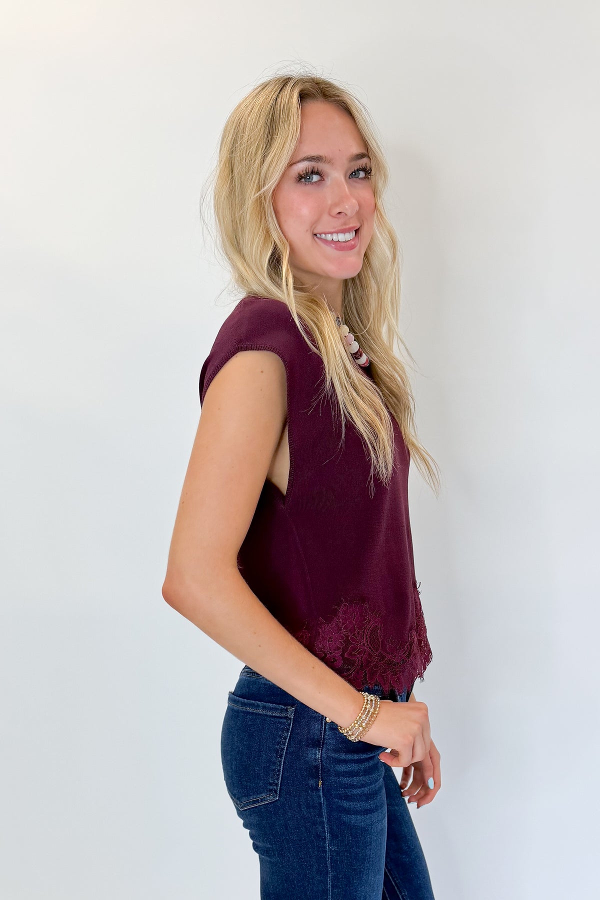 Side view of woman wearing Swagger Boutique’s Wine Short Sleeve Lace Trim Top with jeans against white studio background.