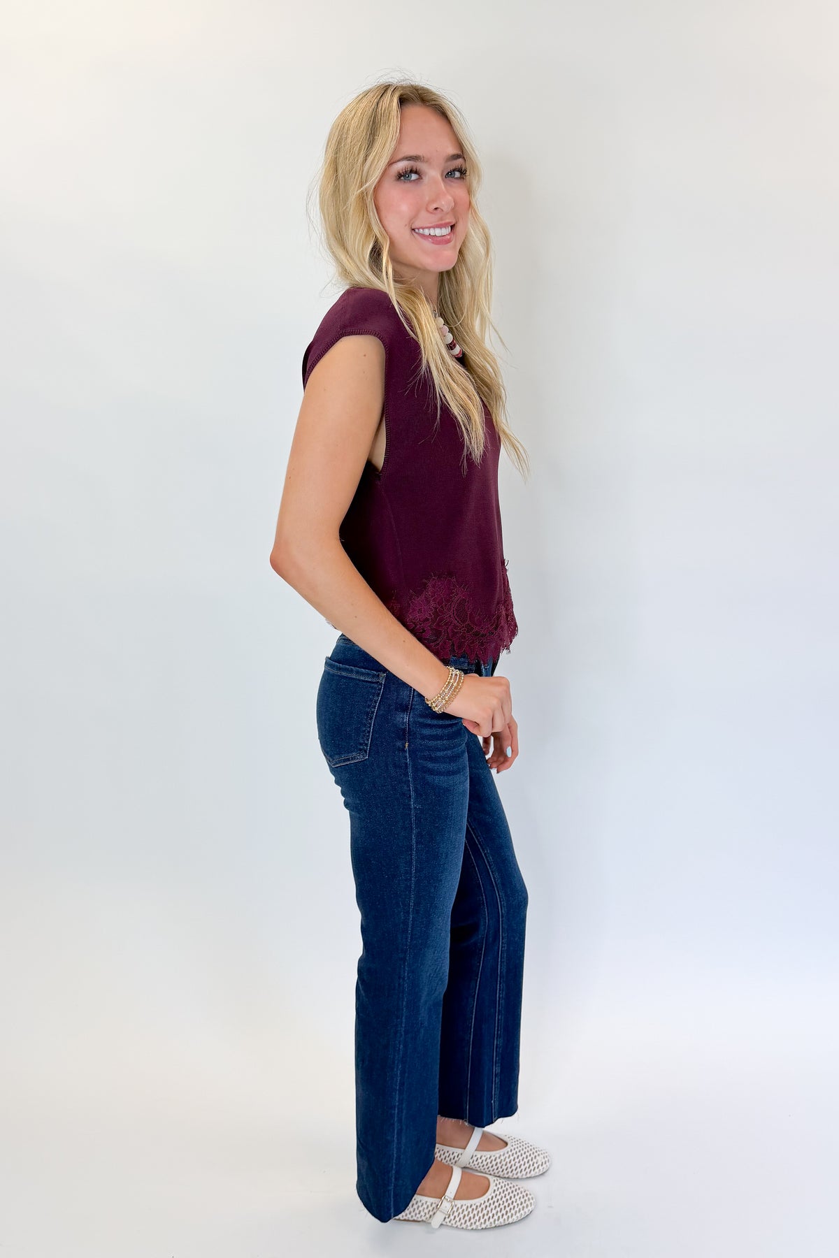 Side view of woman wearing Swagger Boutique’s Wine Short Sleeve Lace Trim Top with jeans against white studio background.