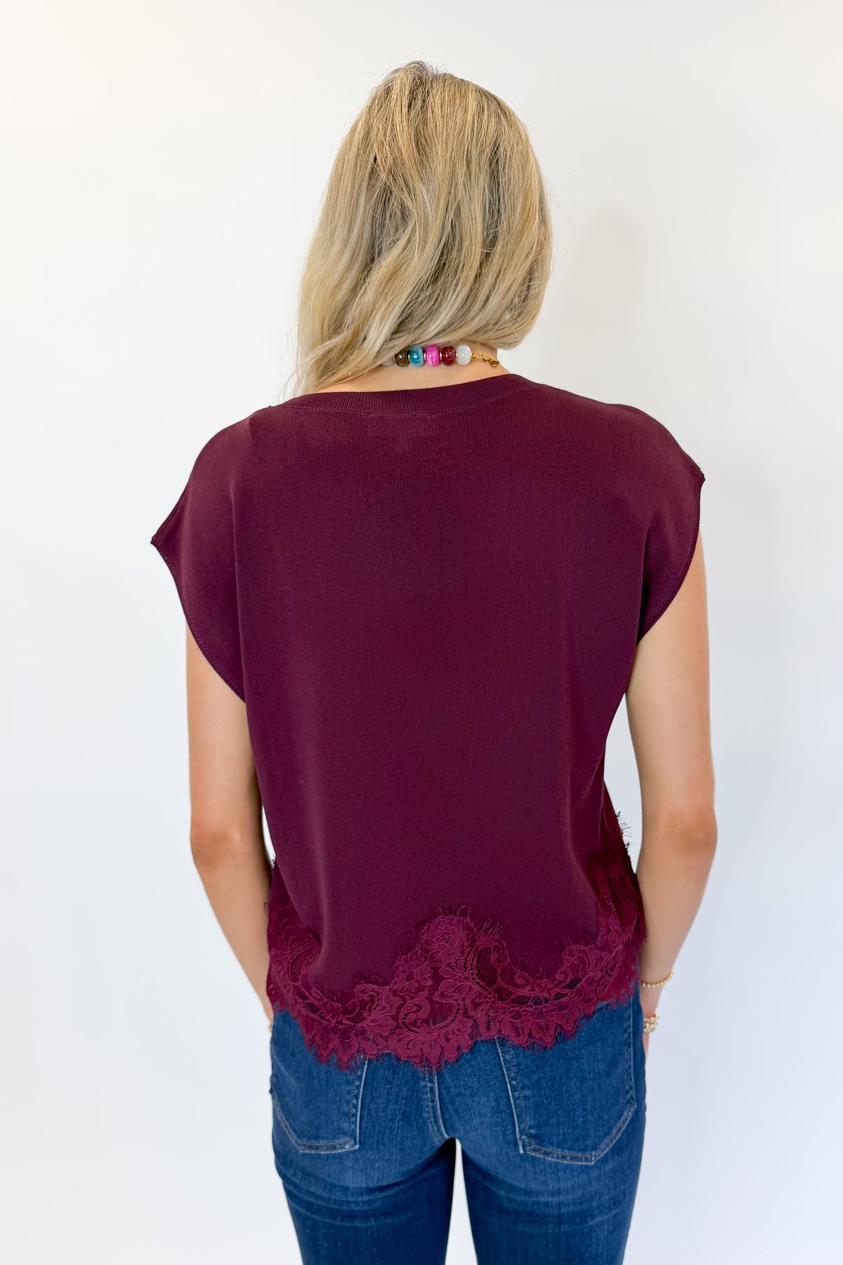 Back view of woman wearing Swagger Boutique’s Wine Short Sleeve Lace Trim Top with jeans against white studio background.