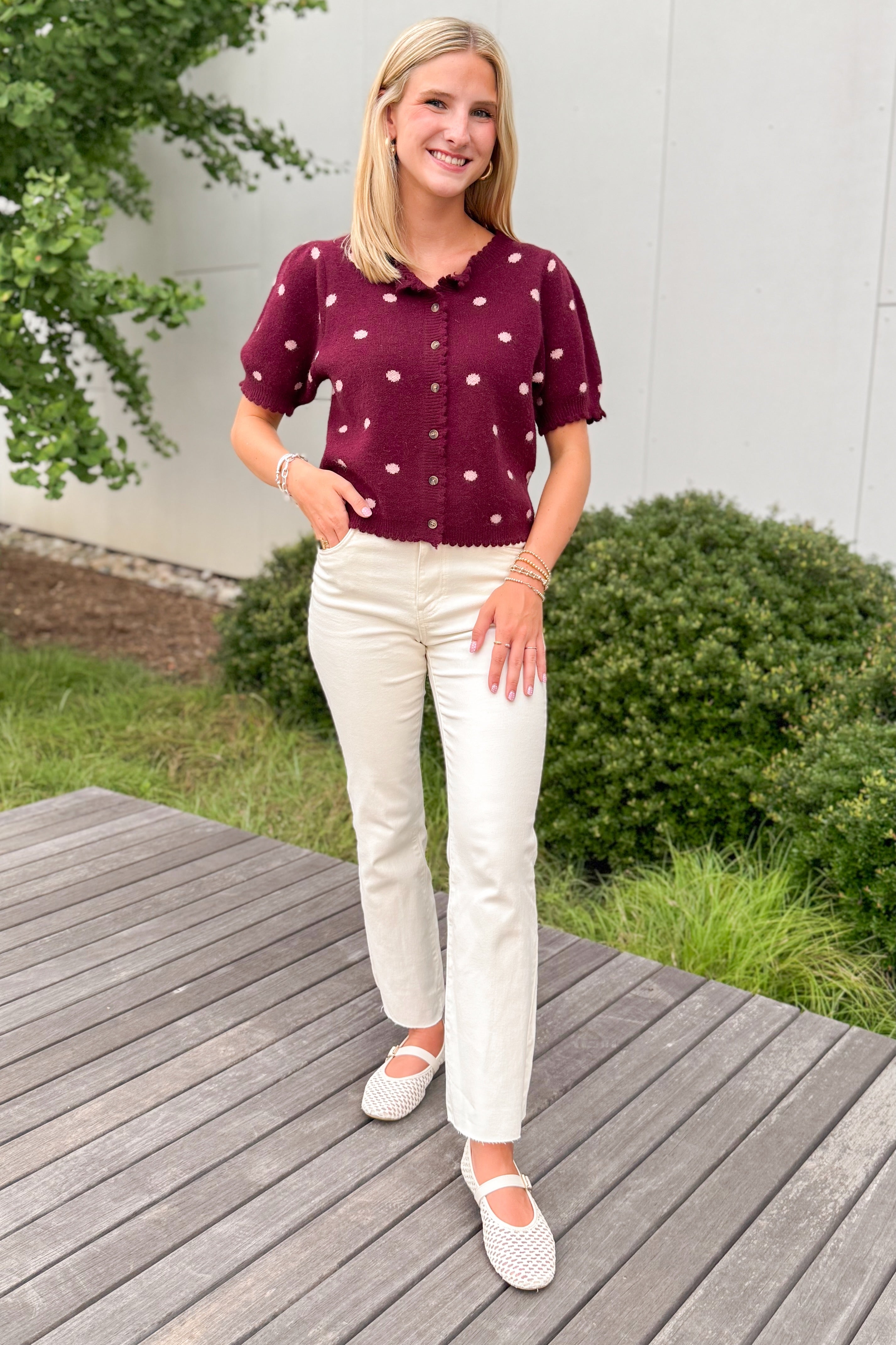 Front view of woman wearing Swagger Boutique’s Wine &amp; Pink Polka Dot Short Sleeve Cardigan by Entro with jeans  standing outside on a deck.