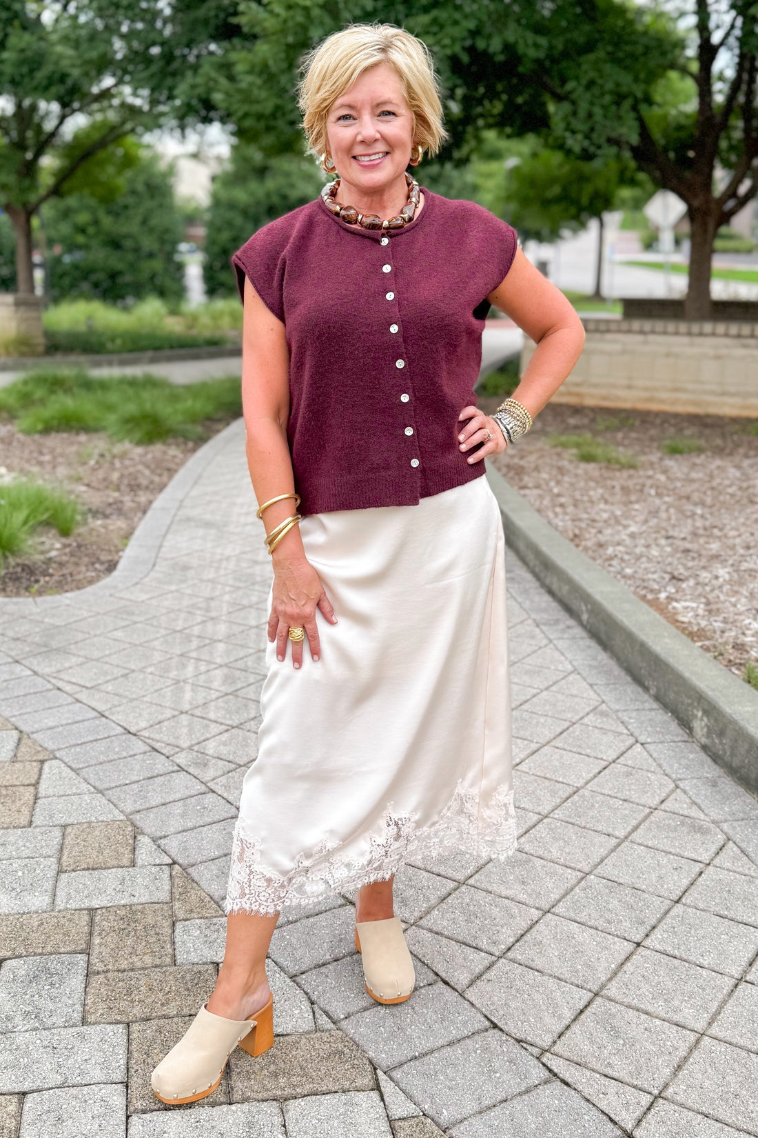 Front view of woman wearing Swagger Boutique’s Wine Sleeveless Button Up Sweater with Ecru Lace Lined Satin Skirt by Entro standing outside at a park.