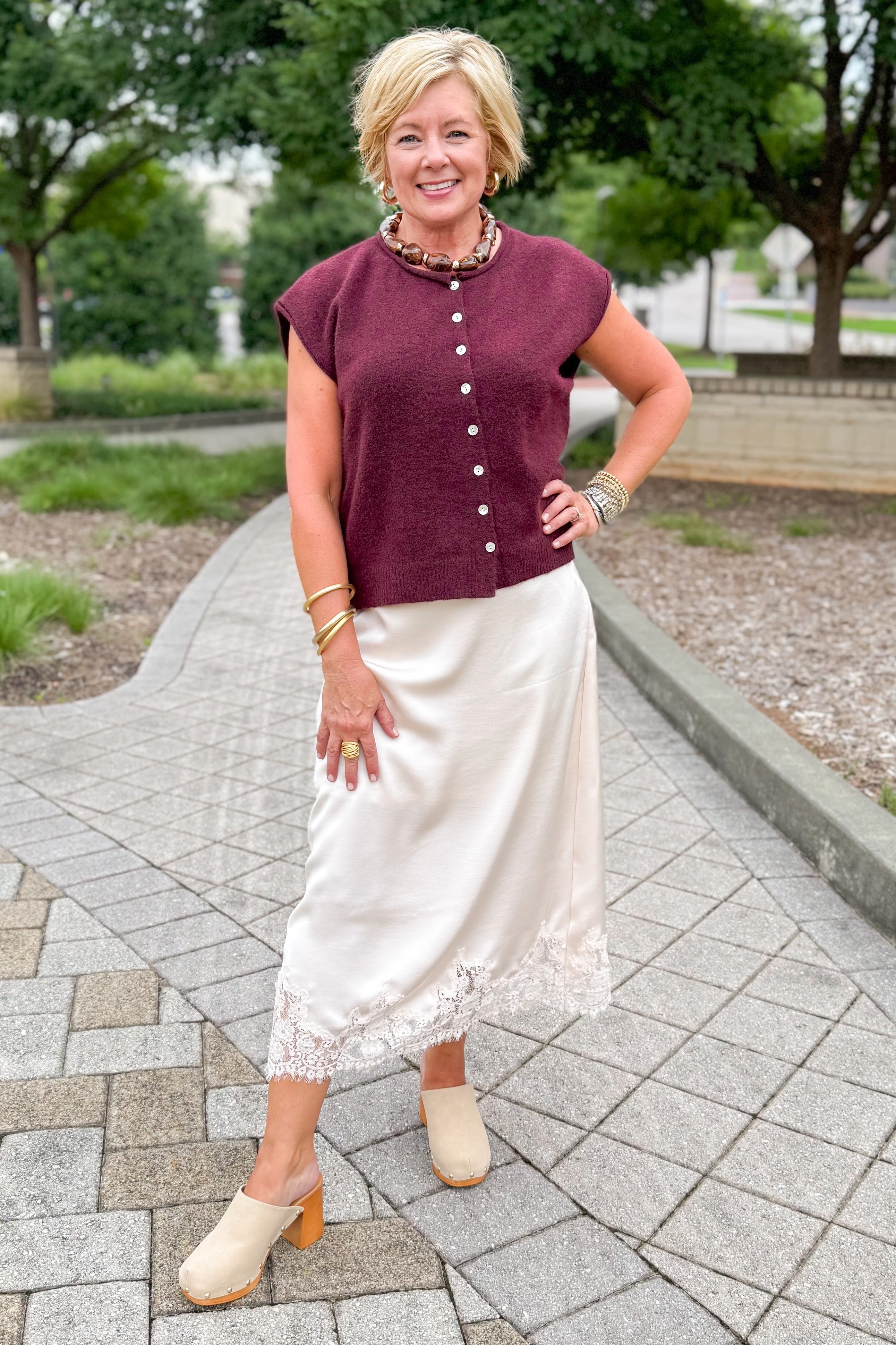 Front view of woman wearing Swagger Boutique’s Wine Sleeveless Button Up Sweater with Ecru Lace Lined Satin Skirt by Entro standing outside at a park.