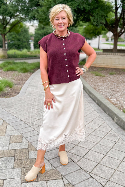 Front view of woman wearing Swagger Boutique’s Wine Sleeveless Button Up Sweater with Ecru Lace Lined Satin Skirt by Entro standing outside at a park.