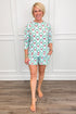 Model wearing the Wreath and Ribbon Annie Pajama Set, featuring green Christmas wreaths with red bows on a light blue and white striped background. The set includes a relaxed-fit, long-sleeve crewneck top and matching pajama shorts with side pockets.