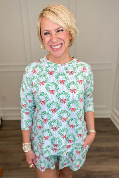 Model wearing the Wreath and Ribbon Annie Pajama Set, featuring green Christmas wreaths with red bows on a light blue and white striped background. The set includes a relaxed-fit, long-sleeve crewneck top and matching pajama shorts with side pockets.