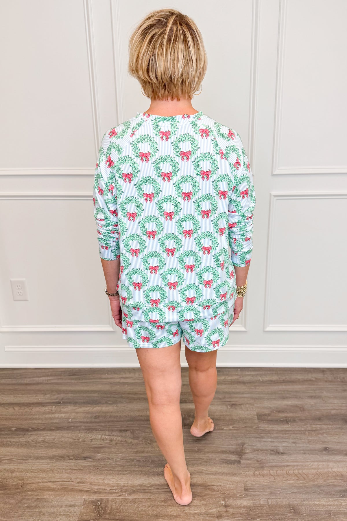 Back view of the Wreath and Ribbon Annie Pajama Set, showcasing the allover green Christmas wreath and red ribbon print against a light blue and white striped background. The long-sleeve pajama top features a comfortable, relaxed silhouette paired with matching shorts.