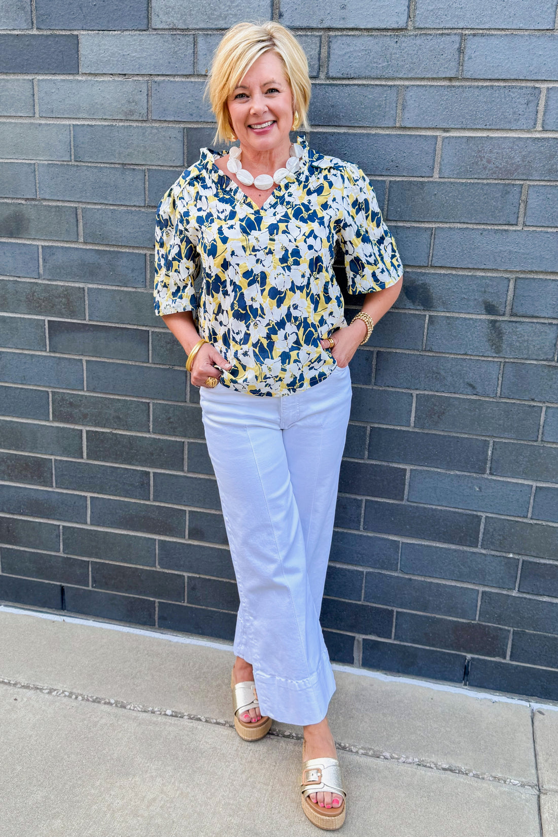 Front view of woman wearing Swagger Boutique’s Yellow &amp; Navy Floral Printed Top with jeans against brick wall outside.