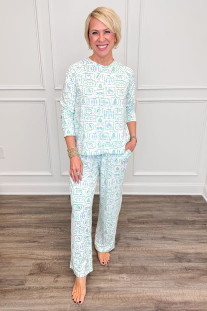 Model wearing the Yueltide Vignettes Blue Annie Pajama Set, featuring a festive light blue and green holiday print on a white background with Christmas trees, wreaths, and decorative holiday motifs. The set includes a relaxed-fit, long-sleeve crewneck top and matching straight-leg pajama pants.