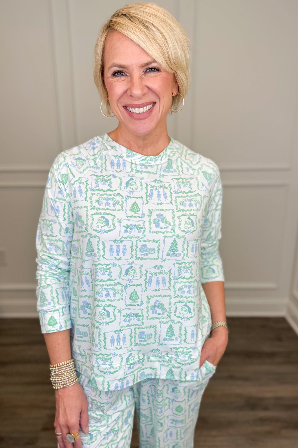 Model wearing the Yueltide Vignettes Blue Annie Pajama Set, featuring a festive light blue and green holiday print on a white background with Christmas trees, wreaths, and decorative holiday motifs. The set includes a relaxed-fit, long-sleeve crewneck top and matching straight-leg pajama pants.