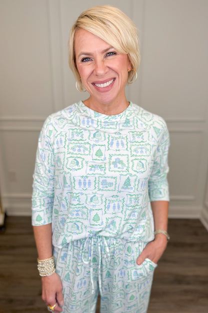 Model wearing the Yueltide Vignettes Blue Annie Pajama Set, featuring a festive light blue and green holiday print on a white background with Christmas trees, wreaths, and decorative holiday motifs. The set includes a relaxed-fit, long-sleeve crewneck top and matching straight-leg pajama pants.