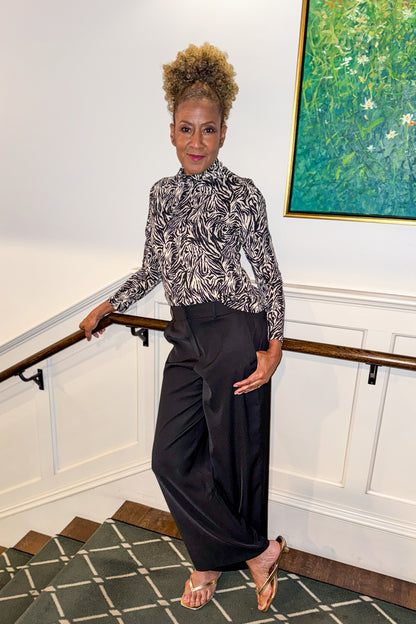 Front view of woman wearing Swagger Boutique’s Black High Waisted Trousers by Skies Are Blue &amp; Zebra Mock Neck Long Sleeve Top by Entro standing by a staircase.