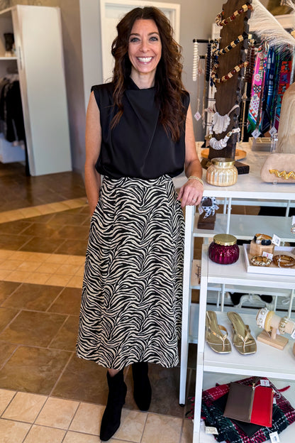 Front view of woman wearing Swagger Boutique’s Zebra Print Midi Skirt by Entro &amp; Black Satin Cowlneck Sleeveless Top  standing at the store.