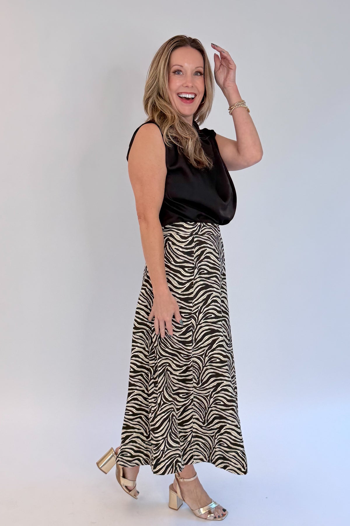 Side view of woman wearing Swagger Boutique’s Zebra Print Midi Skirt by Entro &amp; Black Satin Cowlneck Sleeveless Top  against white studio background.