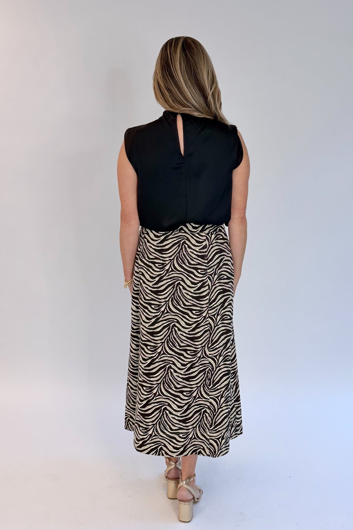 Back view of woman wearing Swagger Boutique’s Zebra Print Midi Skirt by Entro &amp; Black Satin Cowlneck Sleeveless Top  against white studio background.