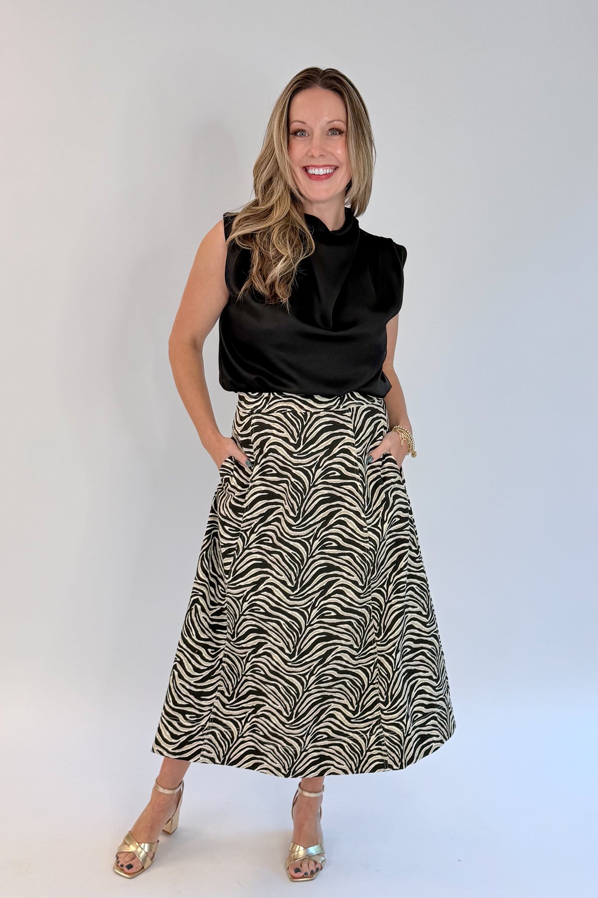 Front view of woman wearing Swagger Boutique’s Zebra Print Midi Skirt by Entro &amp; Black Satin Cowlneck Sleeveless Top  against white studio background.