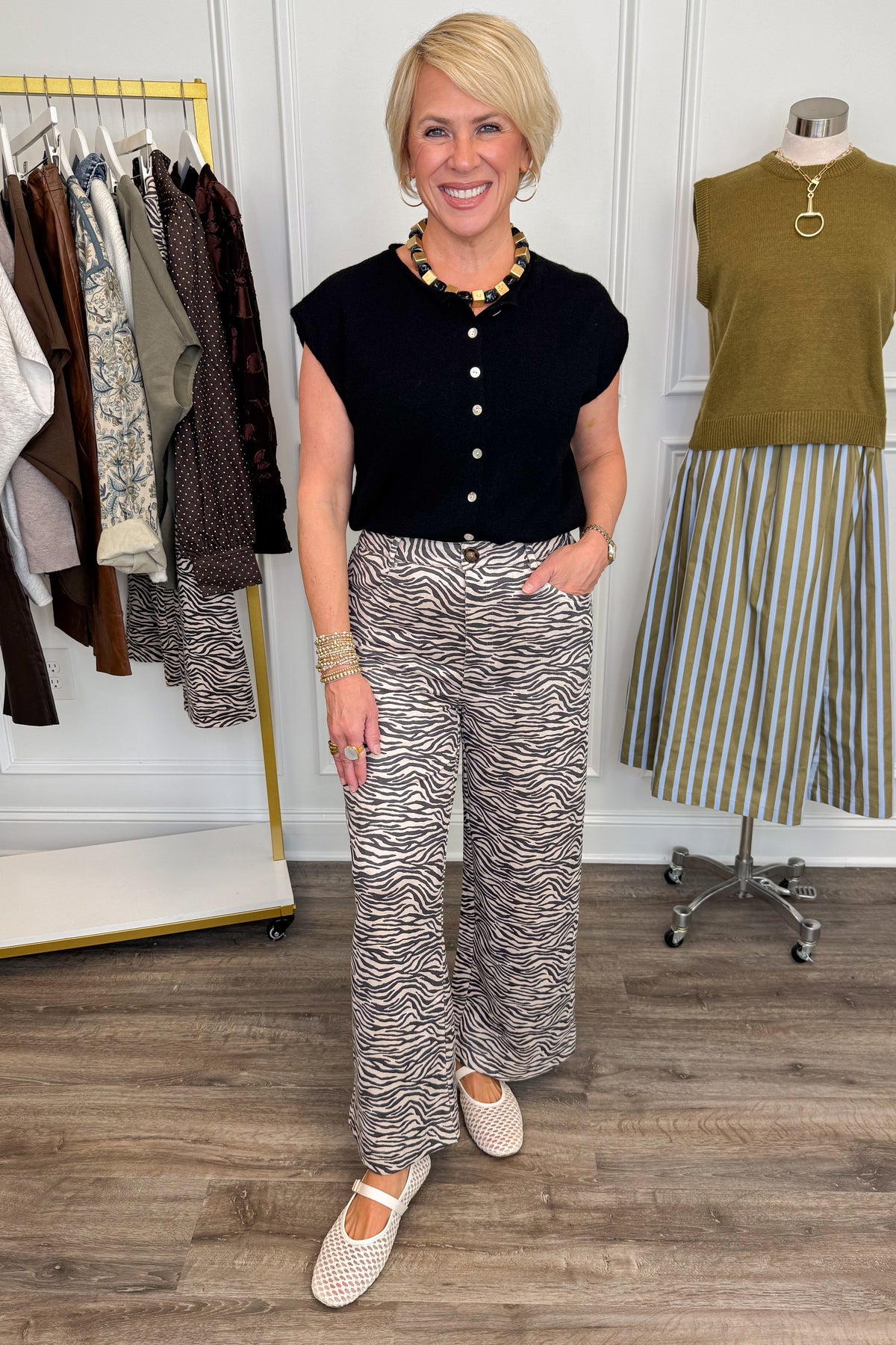 Front view of woman wearing Swagger Boutique’s Zebra Print Suede Pants with Black Sweater with clothing hanging in the background.
