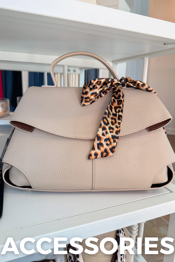 A beige pebbled leather handbag with a top handle and a leopard-print ribbon tied in a bow, displayed on a white shelf. The word 'ACCESSORIES' is overlaid at the bottom.