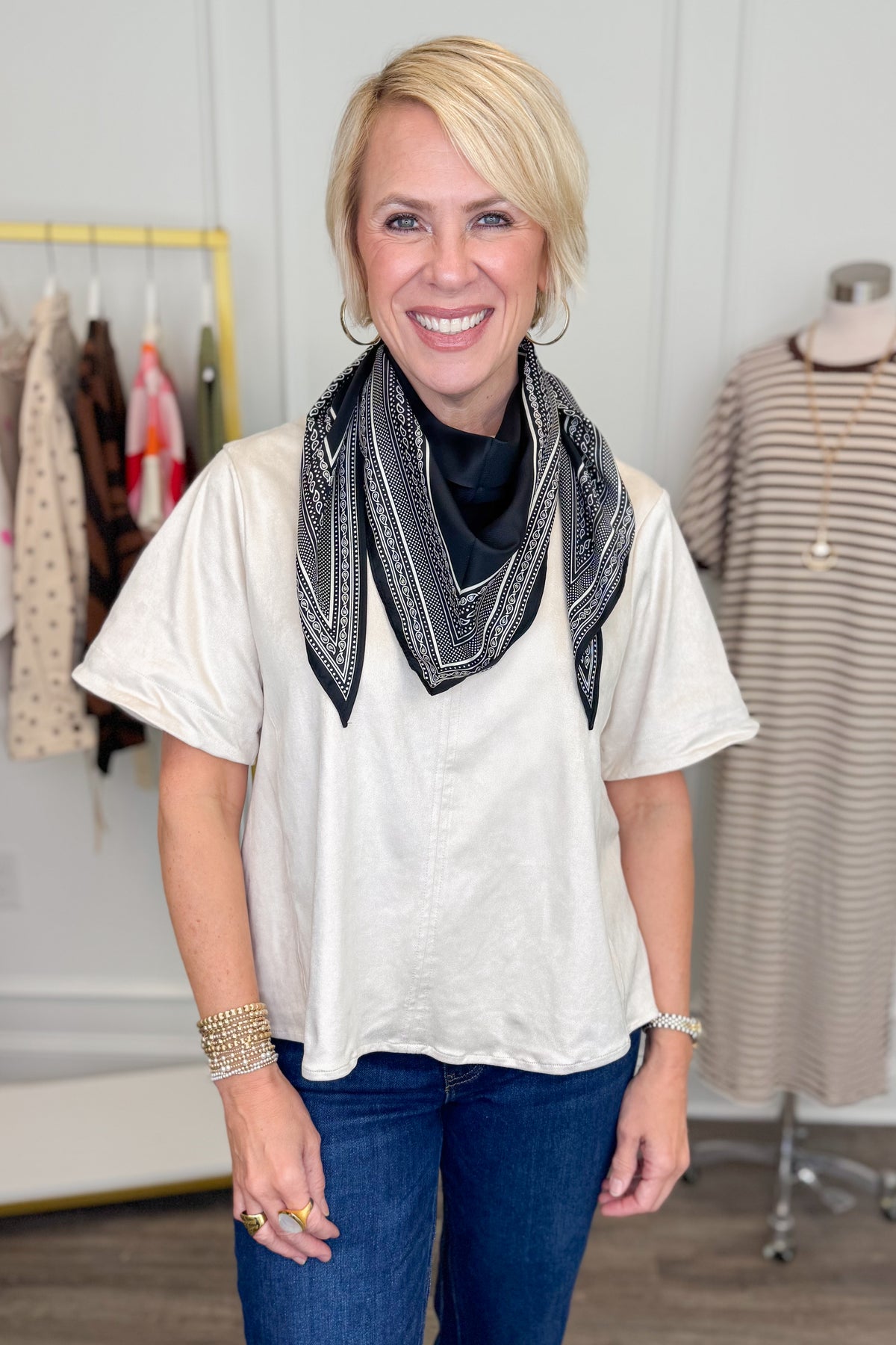 A smiling blonde woman wearing a cream short-sleeve top, blue jeans, and a black bandana-style scarf standing in a boutique setting.