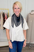 A smiling blonde woman wearing a cream short-sleeve top, blue jeans, and a black bandana-style scarf standing in a boutique setting.