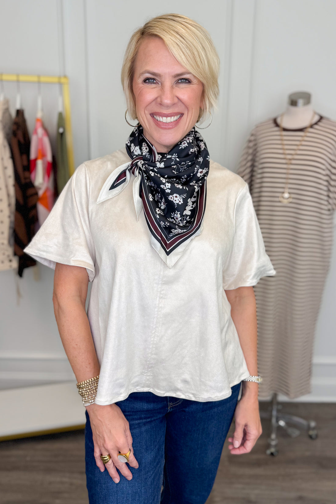 A smiling blonde woman stands indoors wearing a loose short-sleeve cream blouse and blue jeans, with a black floral neck scarf. A clothing rack and a dress form are visible in the boutique setting behind her.