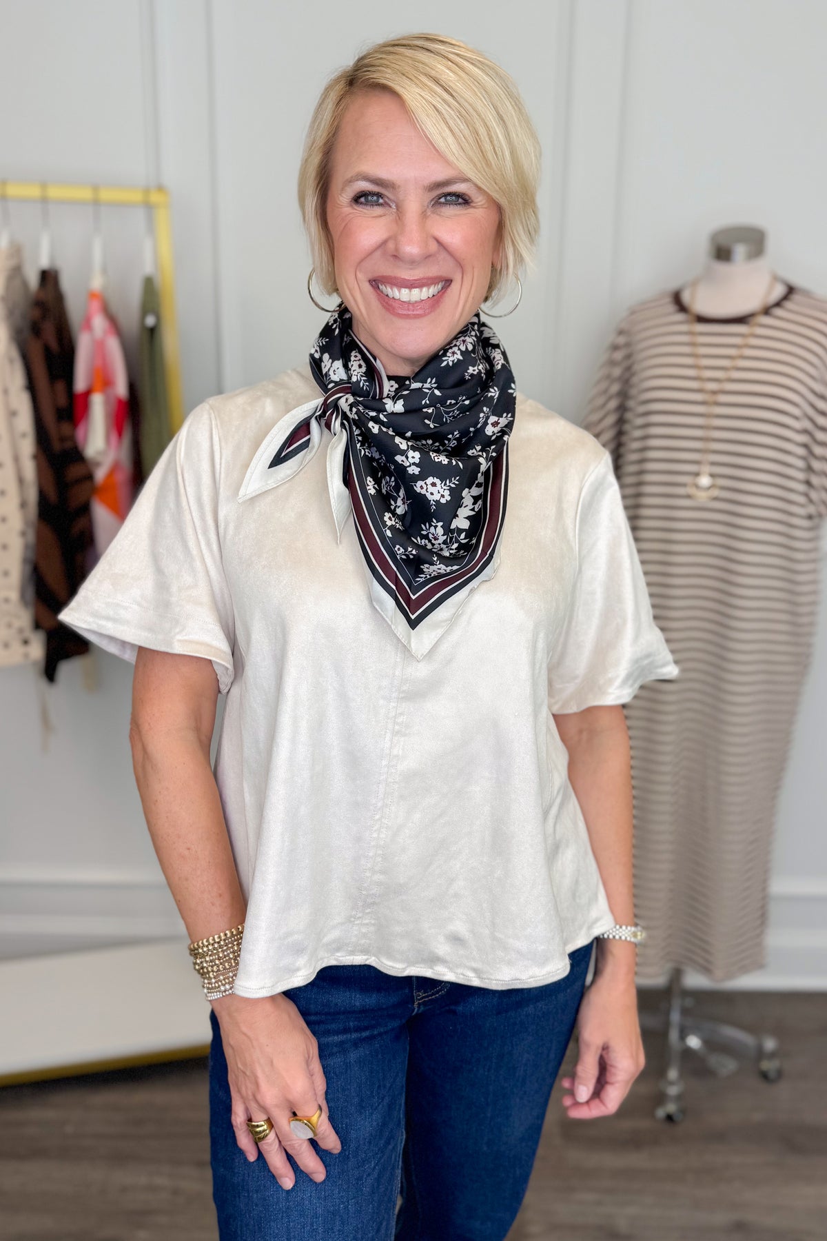 A smiling blonde woman stands indoors wearing a loose short-sleeve cream blouse and blue jeans, with a black floral neck scarf. A clothing rack and a dress form are visible in the boutique setting behind her.