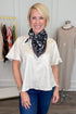 A smiling blonde woman stands indoors wearing a loose short-sleeve cream blouse and blue jeans, with a black floral neck scarf. A clothing rack and a dress form are visible in the boutique setting behind her.