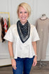 A smiling blonde woman wearing a cream short-sleeve top and blue jeans, styled with a black and white paisley bandana scarf, standing in a boutique setting.