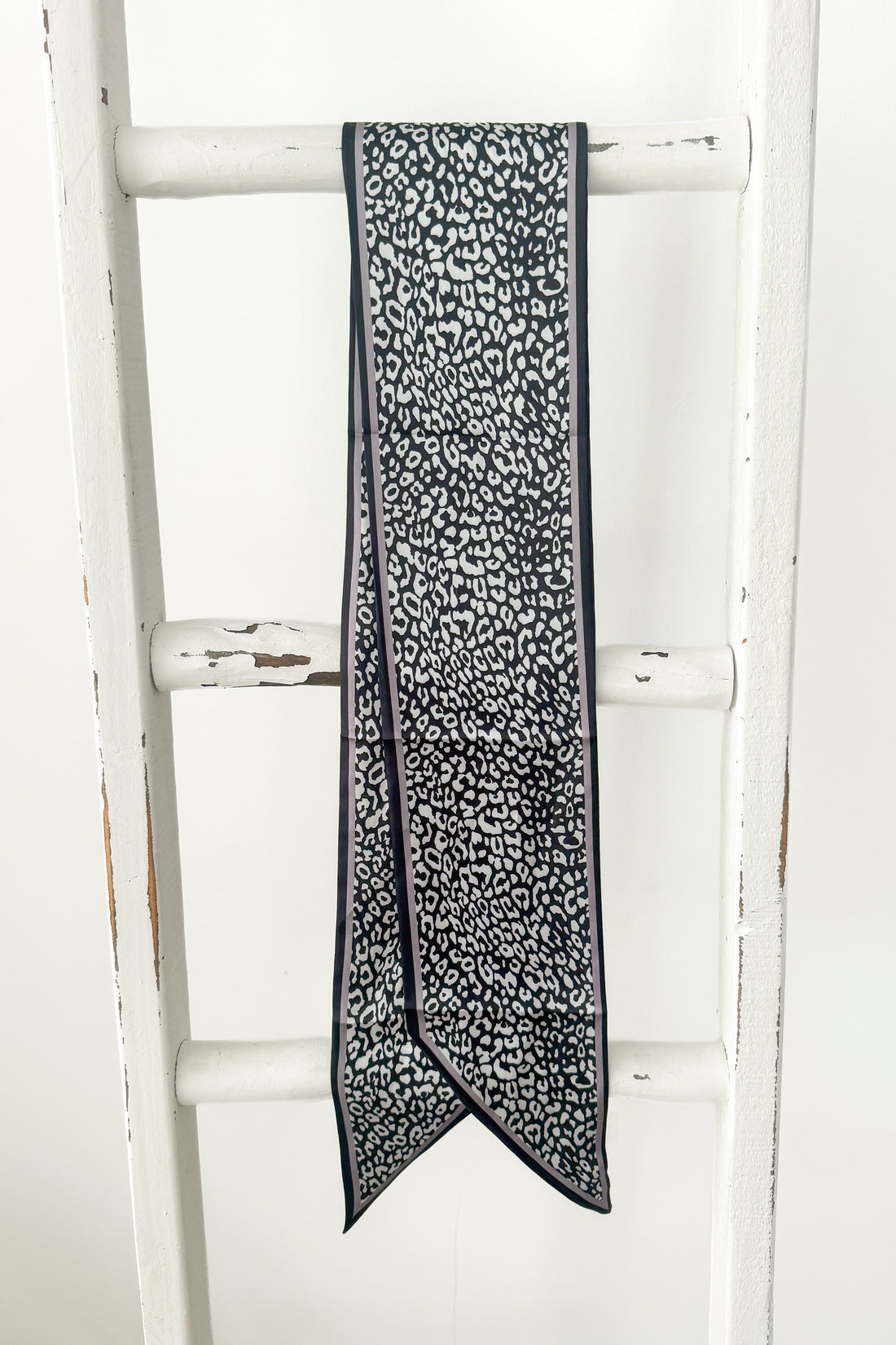 A long narrow scarf with a black and white leopard print pattern, draped over the rung of a white wooden ladder.