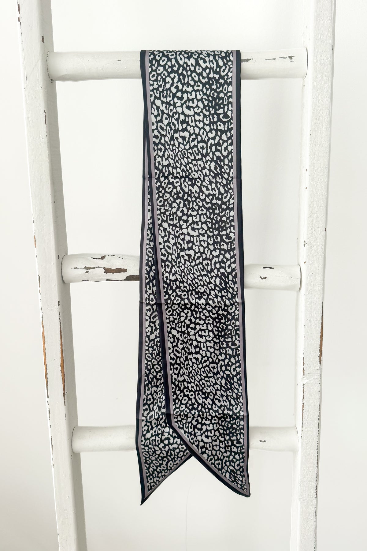 A long narrow scarf with a black and white leopard print pattern, draped over the rung of a white wooden ladder.