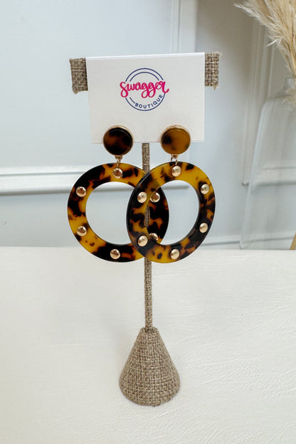 Tortoiseshell hoop earrings with gold-tone studs, displayed on a burlap stand with a white card labeled &