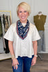 A smiling blonde woman stands in a boutique wearing a cream short-sleeve button-up top, blue jeans, and a navy floral neck scarf with red and white stripes. A clothing rack and a mannequin are visible in the softly lit store interior.
