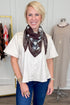 A smiling blonde woman wearing a cream short-sleeve top, blue jeans, and a brown bandana-style scarf with a white and gray floral print, standing in a boutique setting.