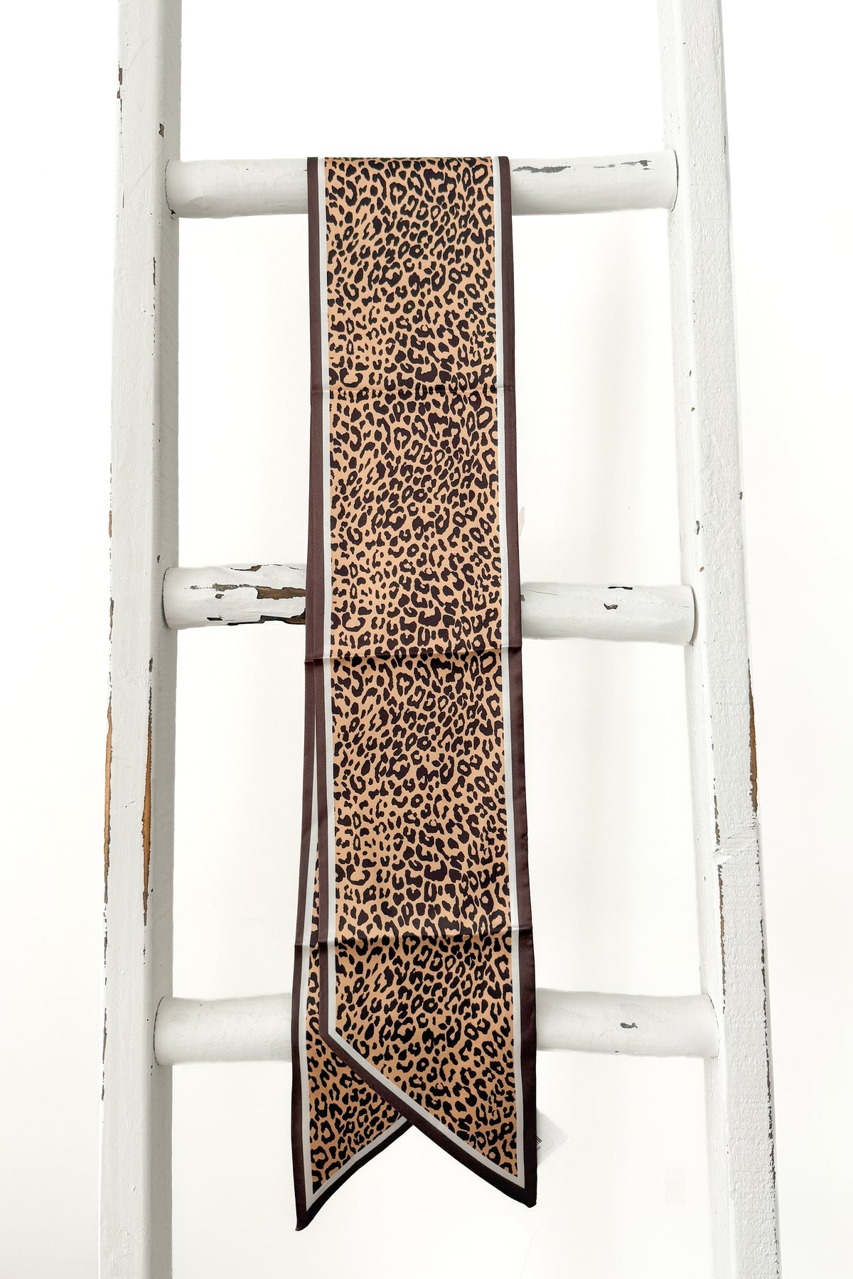 A leopard print scarf with dark brown borders, draped over the rungs of a rustic white wooden ladder.