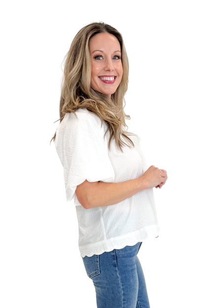 Side view of woman wearing White Scallop Detail V-neck Top with jeans against white studio background.