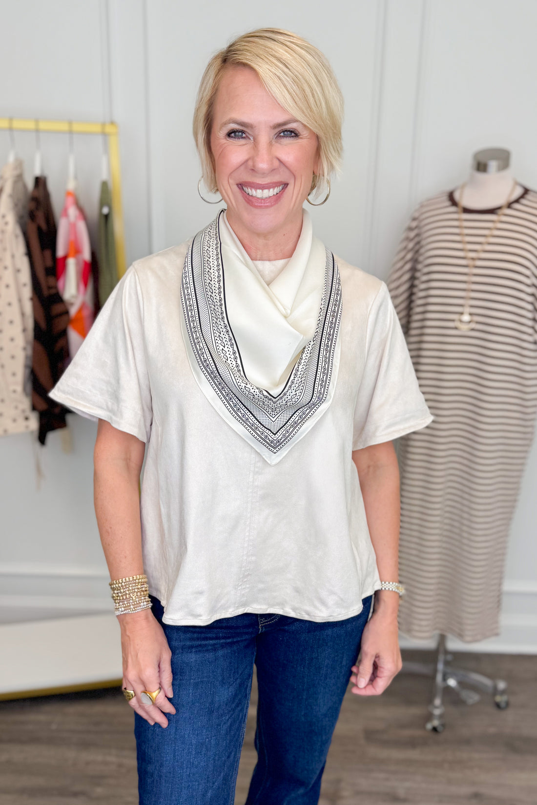 A smiling blonde woman stands indoors wearing a loose short-sleeve cream top with a wide embroidered V-neck trim and blue jeans. A clothing rack and a dress form are visible in the softly lit boutique setting.