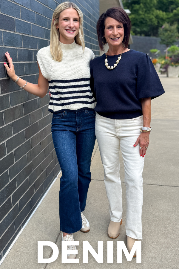 Two women standing outdoors next to a gray brick wall, showcasing denim apparel. The woman on the left wears a cream and navy striped knit sweater vest with dark blue jeans, and the woman on the right wears a navy short-sleeve top with white jeans.