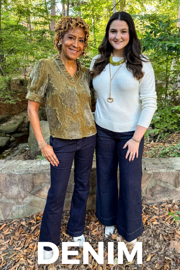 Two women smiling outdoors in a leafy garden, wearing dark-wash denim jeans paired with a textured olive blouse and a white sweater. White text reads “DENIM.”