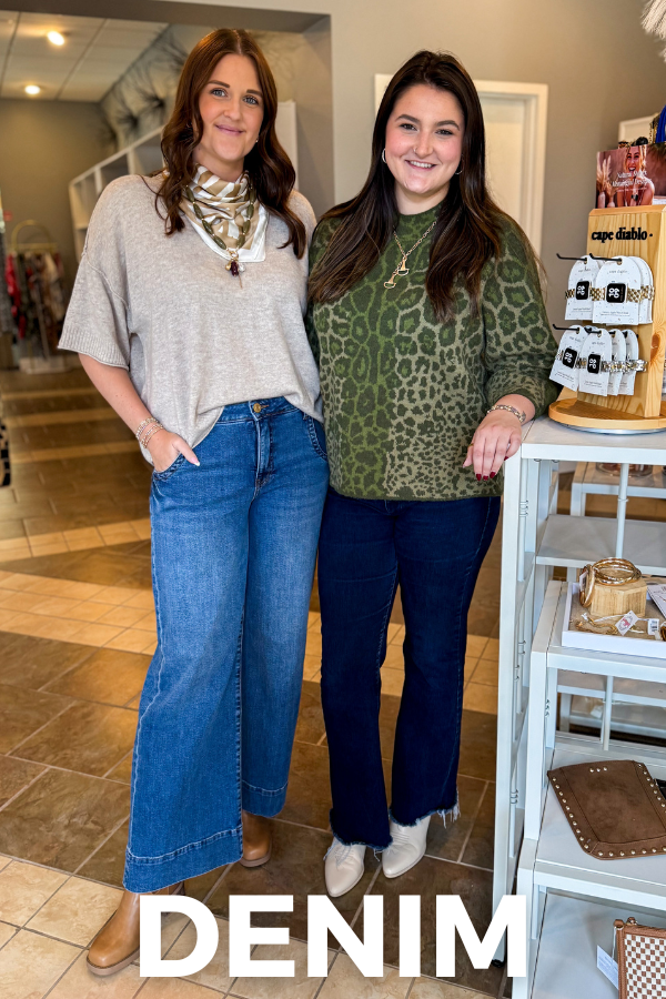 Two women standing in a retail store interior, both wearing blue denim jeans. The woman on the left wears a light gray tunic top and a patterned scarf, while the woman on the right wears a green leopard-print top and dark blue jeans.