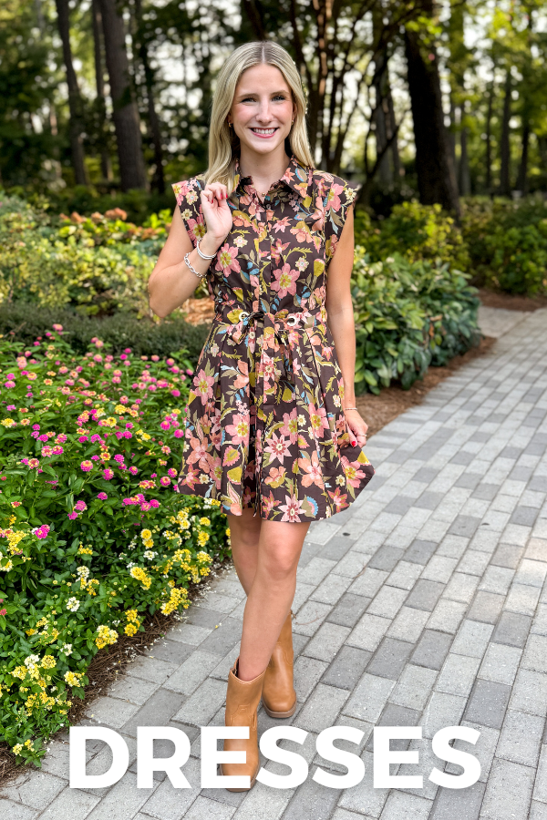 A woman stands on a paved garden path wearing a sleeveless floral mini dress with a button-front bodice and flared skirt. The dress has a dark brown base with pink, yellow, and orange floral print.
