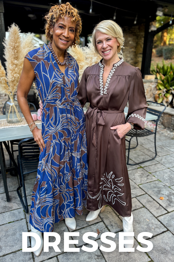 Two women posing together on a stone patio, wearing patterned dresses—one blue with a flowing floral design and the other brown with white decorative stitching—paired with light-colored boots. White text reads “DRESSES.”
