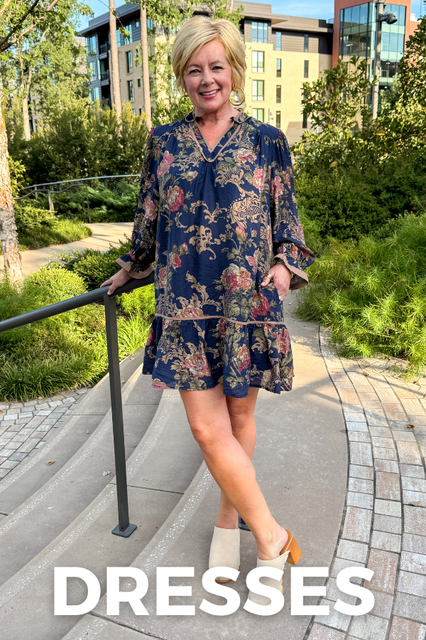 A woman stands on outdoor steps wearing a navy floral mini dress with a tie neckline and ruffled hem. The dress has a multicolor floral pattern with pink, red, and beige tones.