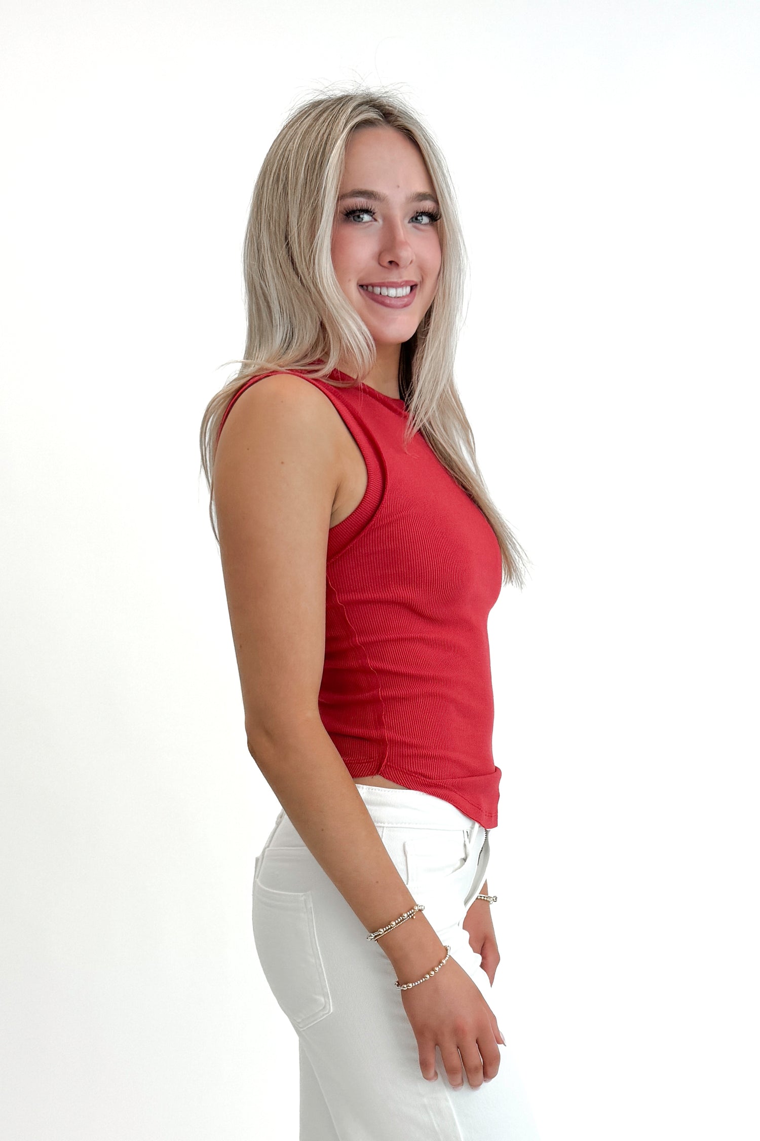 Side view of woman wearing Red Round Hem Ribbed Tank with jeans against white studio background.