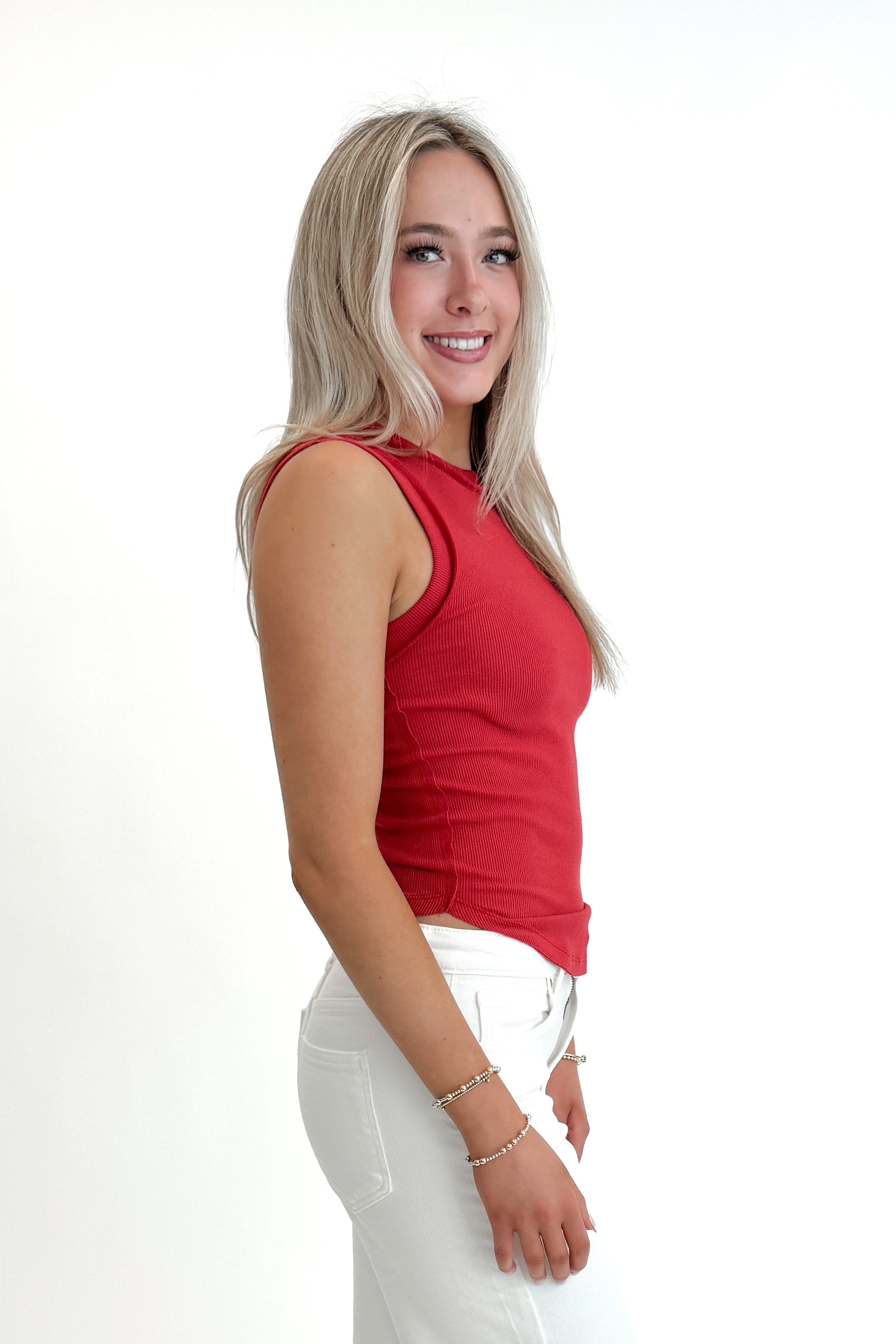 Side view of woman wearing Red Round Hem Ribbed Tank with jeans against white studio background.