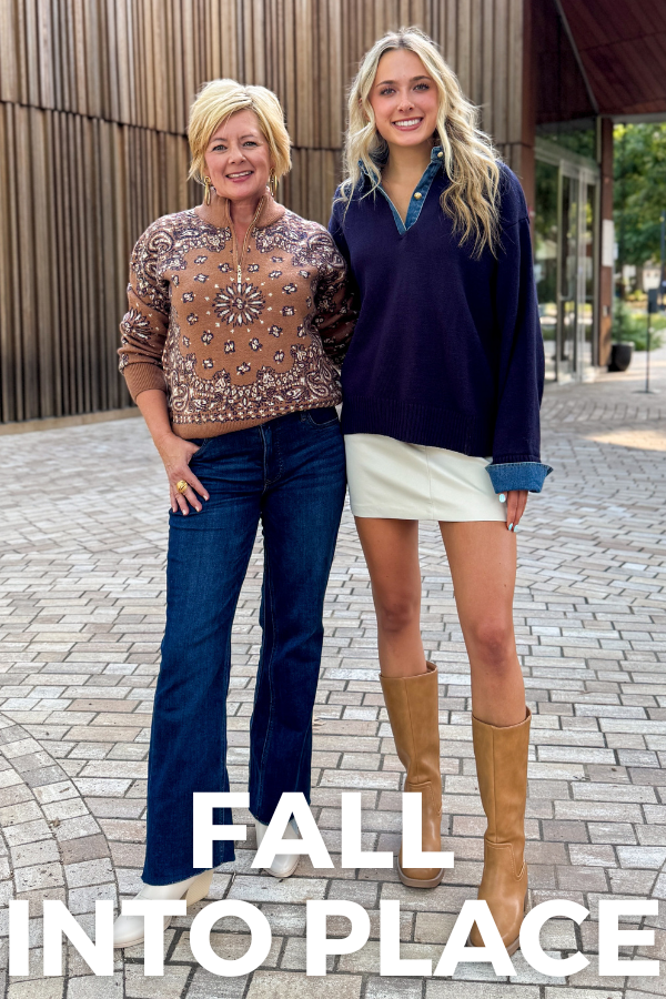 Two women standing outdoors on a paved walkway in front of a modern building with wood siding. The woman on the left wears a brown patterned long-sleeve top and dark blue jeans, while the woman on the right wears a navy quarter-zip pullover, a white mini skirt, and tan knee-high boots.