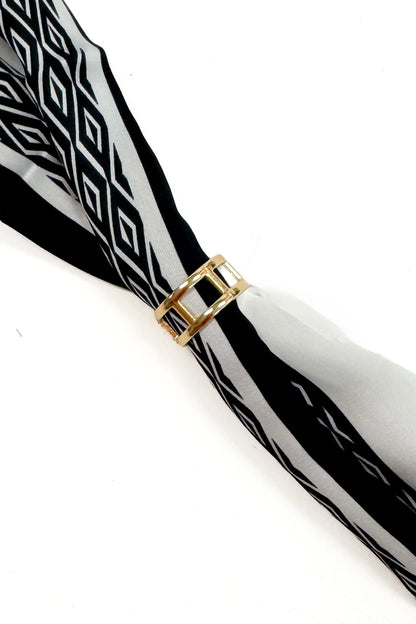 A close-up of a black and white patterned scarf tied with a gold metal ring at the center, set against a plain white background.
