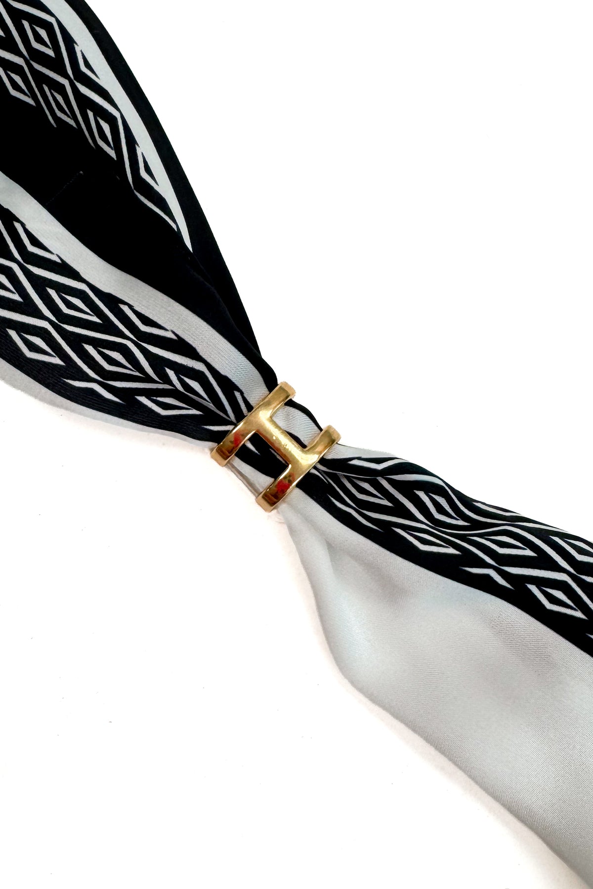 A close-up of a black and white patterned scarf tied with a gold H-shaped ring clasp, set against a plain white background.