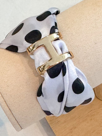 A close-up of a bracelet or cuff featuring a white satin fabric with black polka dots and a prominent polished gold metal H-shaped clasp, displayed on a beige cylindrical stand.