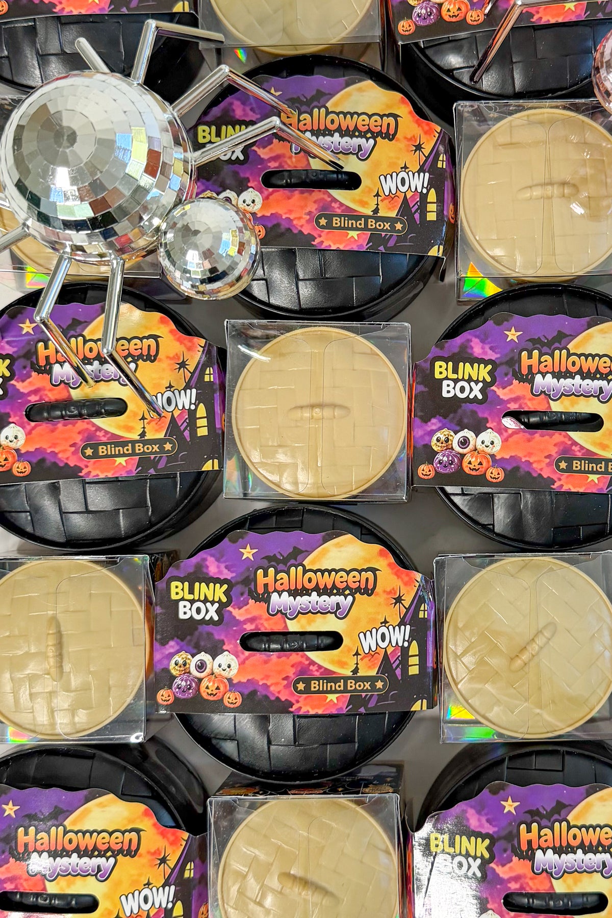 Multiple Halloween mystery blind box products arranged in a grid, each showing a beige fake wound makeup disc in a clear plastic package with a black basket base and a colorful Halloween-themed label. A disco ball prop is visible in the upper left.