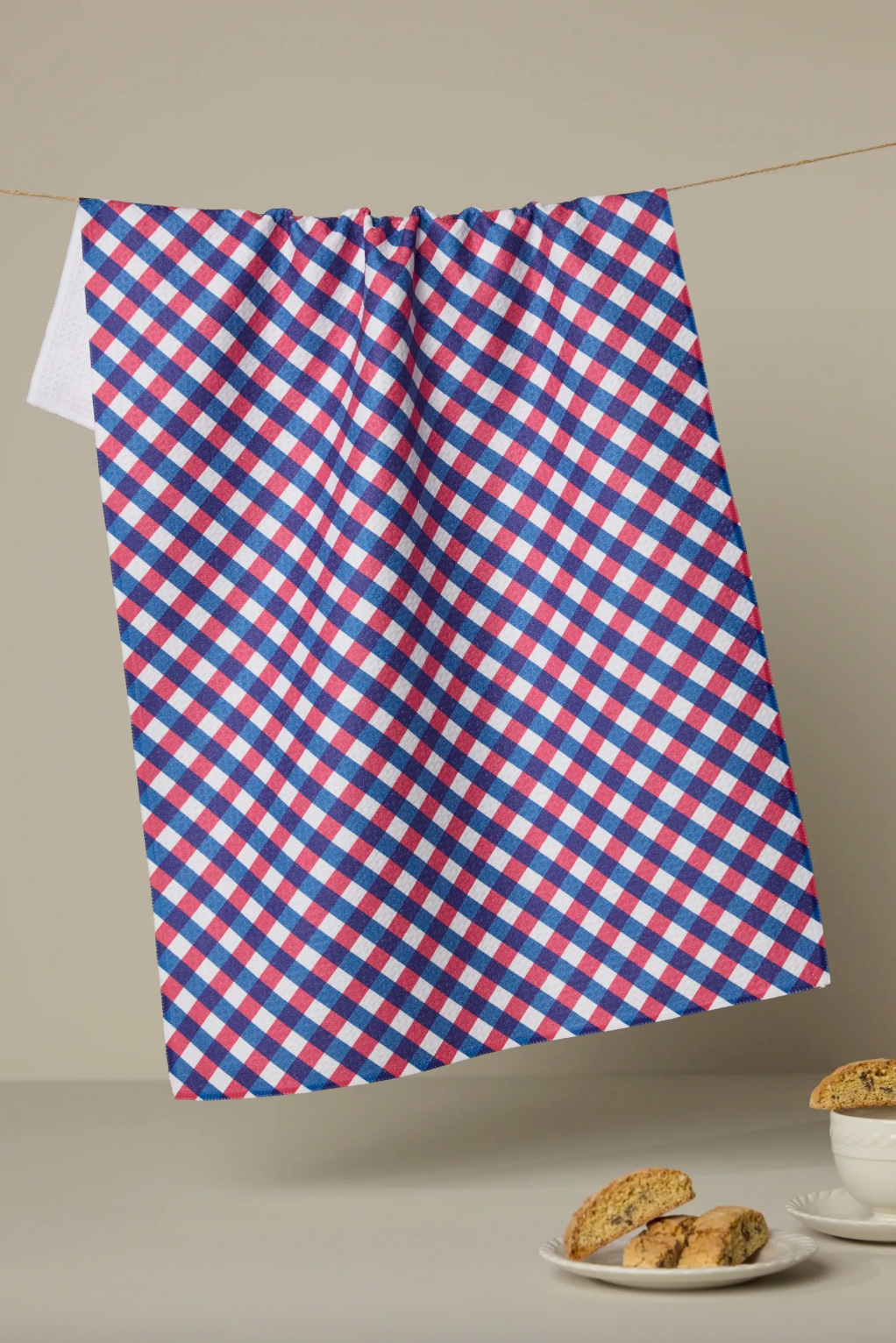 Red, white, and blue checkered towel hanging on a line with cookies and a cup on a table below.