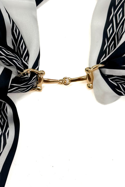 A close-up of a black and white patterned scarf tied with a gold horsebit clasp on a white background.
